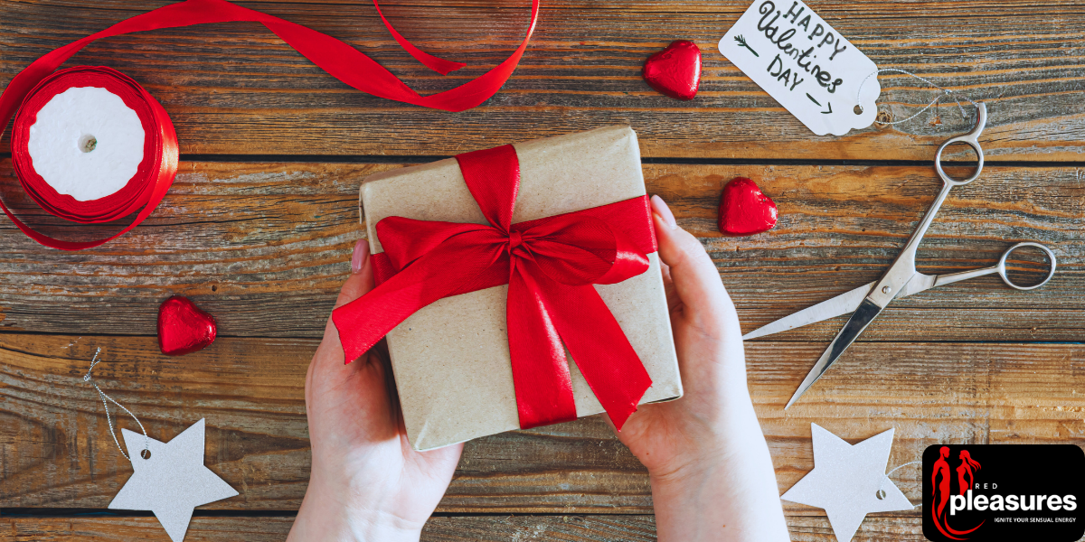 Hands holding a small gift wrapped with a red ribbon over a table scattered with hearts and craft supplies.