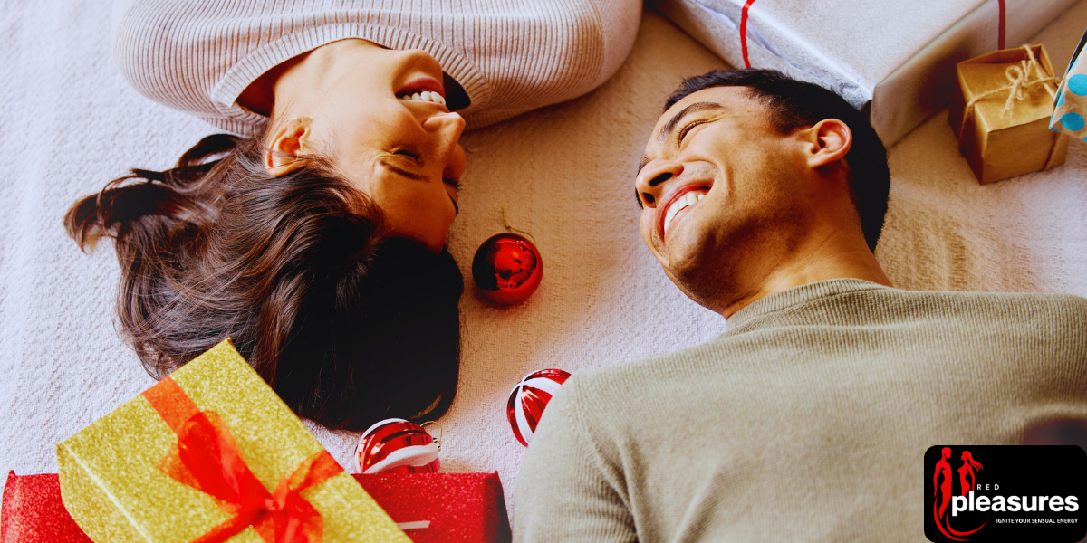 A smiling couple lying on a bed surrounded by Valentine's gifts and red hearts.