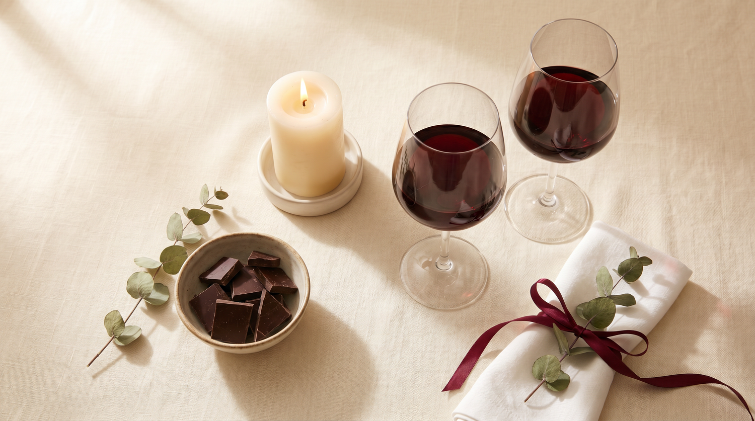 Romantic at-home date night setup with red wine, candle, dark chocolate, and a linen napkin tied with a burgundy ribbon