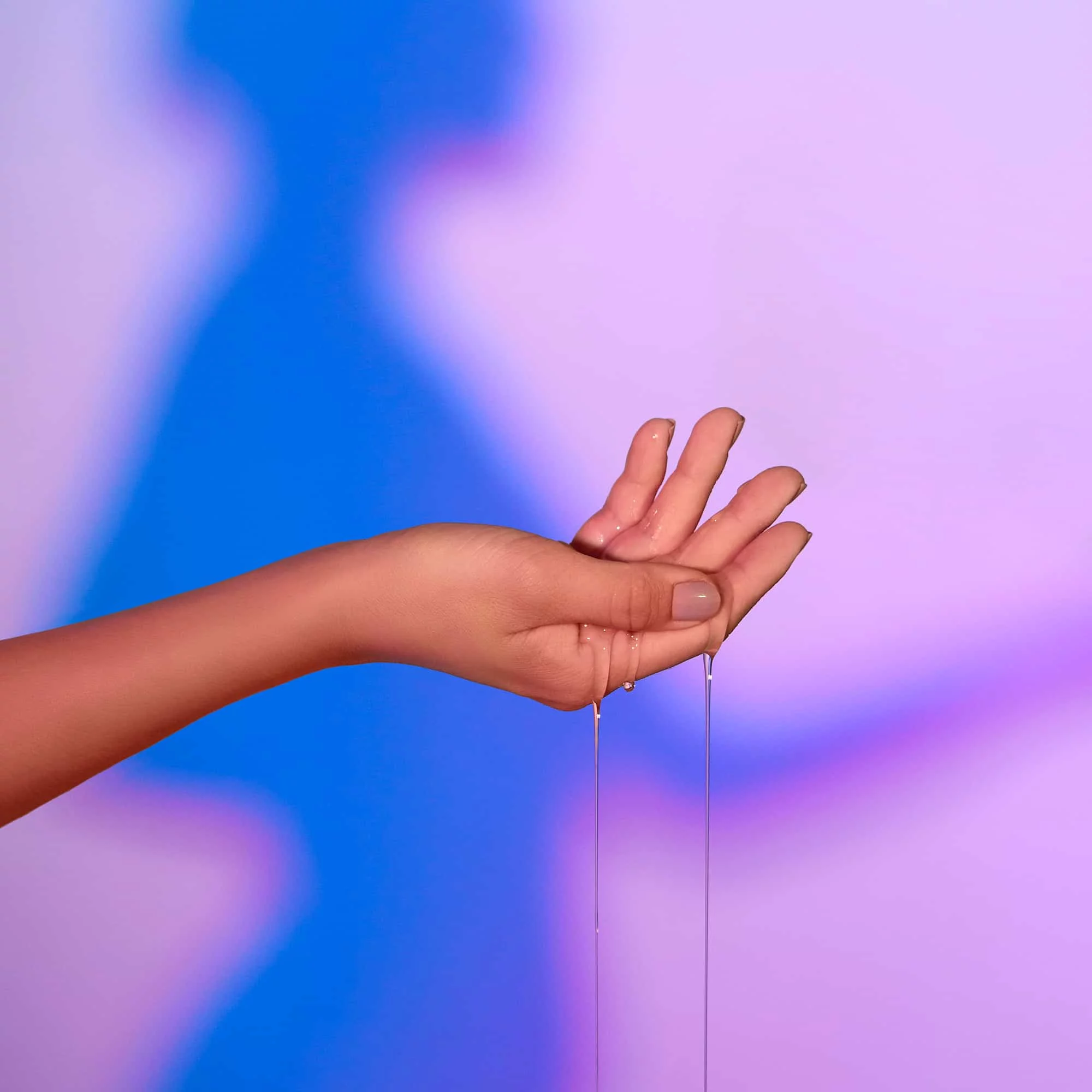 A hand holds warming massage oil that drips down against a blurred blue and pink background.