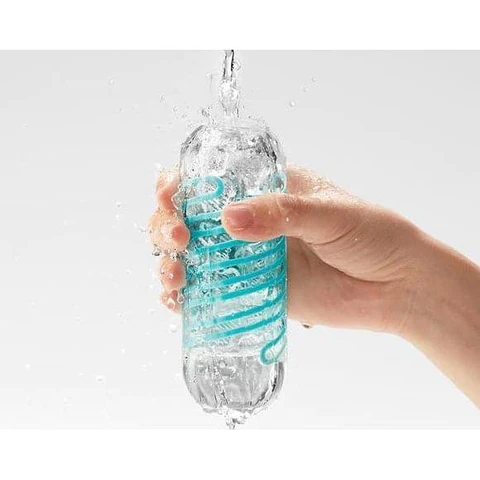A hand holds a clear Tenga Spinner 01 Tetra masturbator with a blue spiral inside under running water.