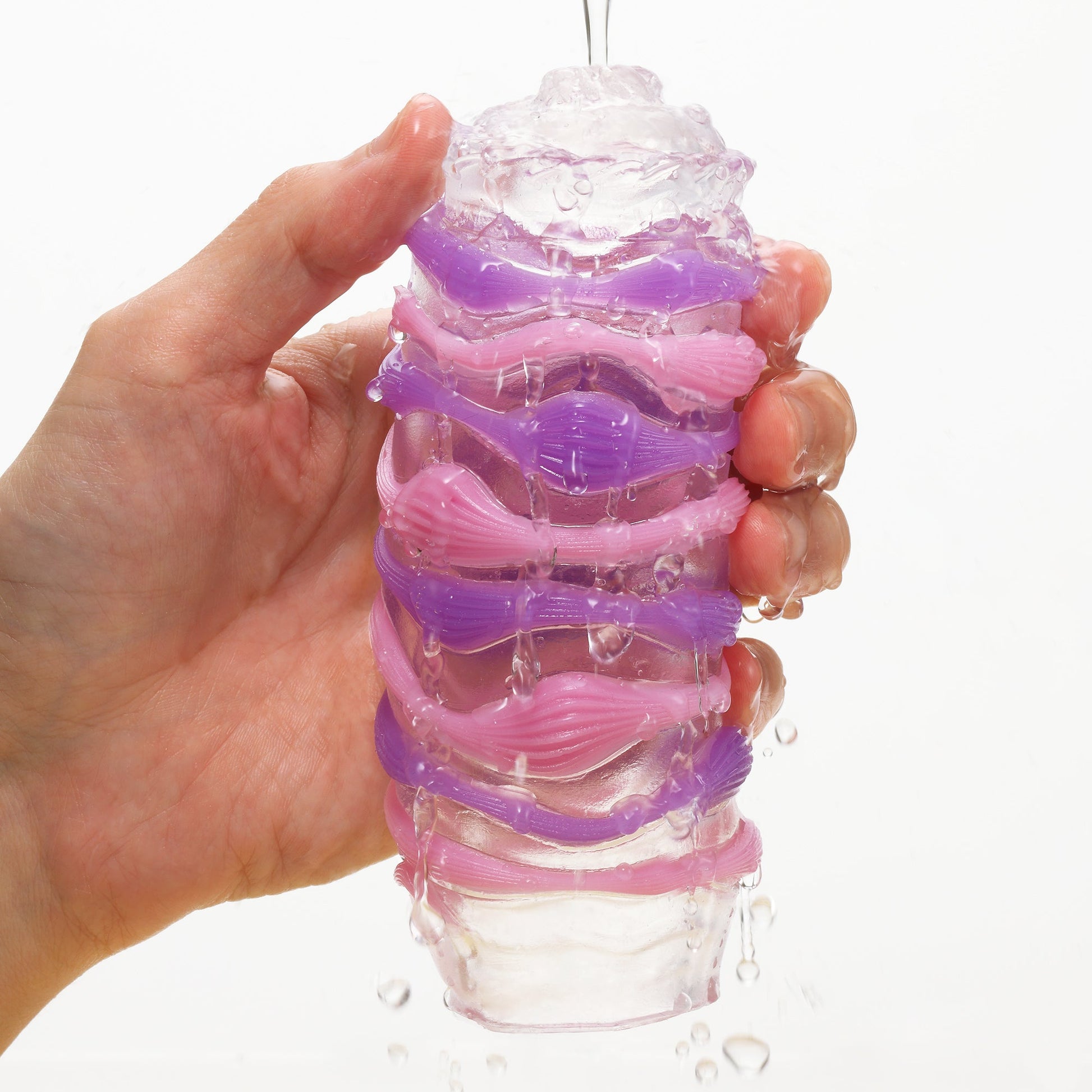 A hand holds a clear, textured male masturbator with pink and purple ribbed layers.