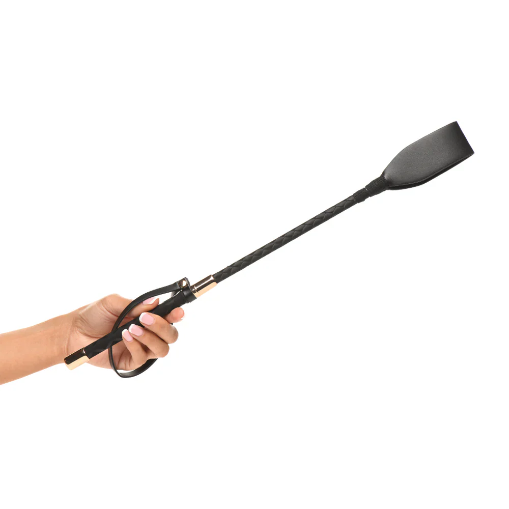 A hand holds a black vegan leather riding crop with a textured grip and gold accents.