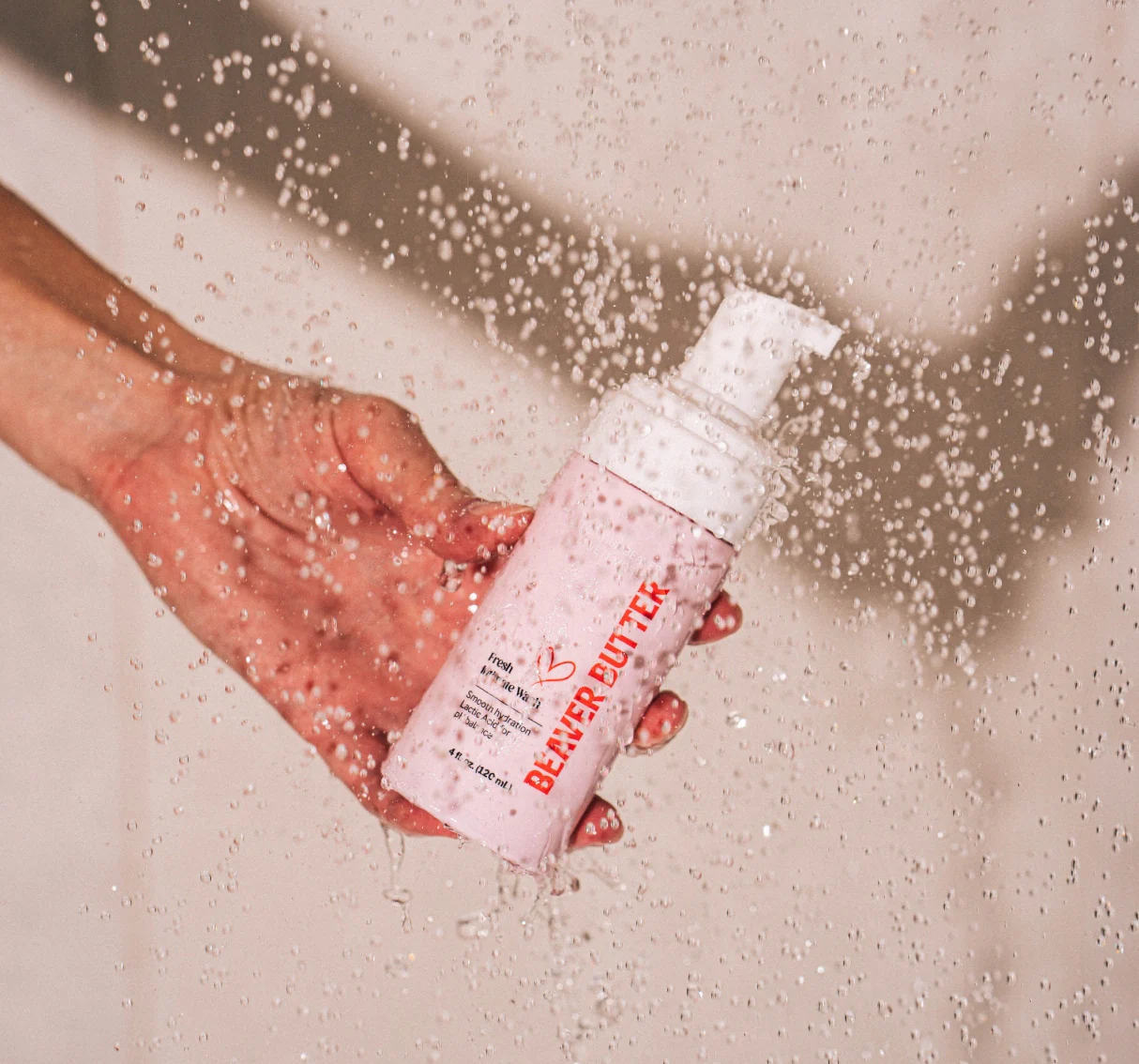 A hand holds a pink bottle of Beaver Butter intimate wash under a shower spray.