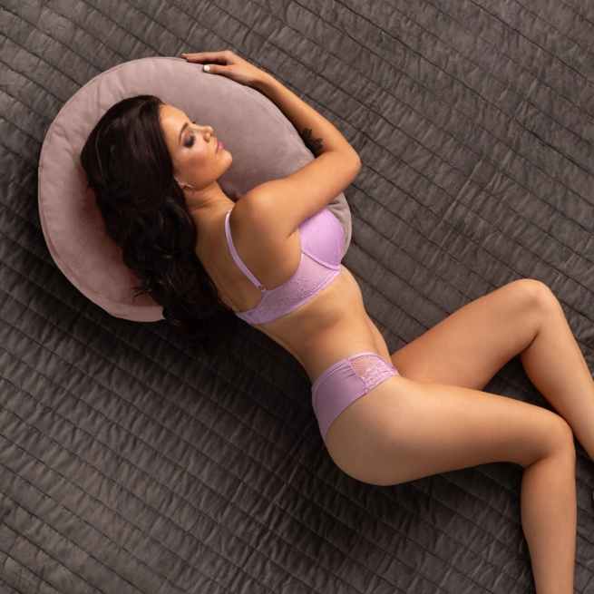 A woman in a purple bra and underwear lies on a gray bed with a round pillow.