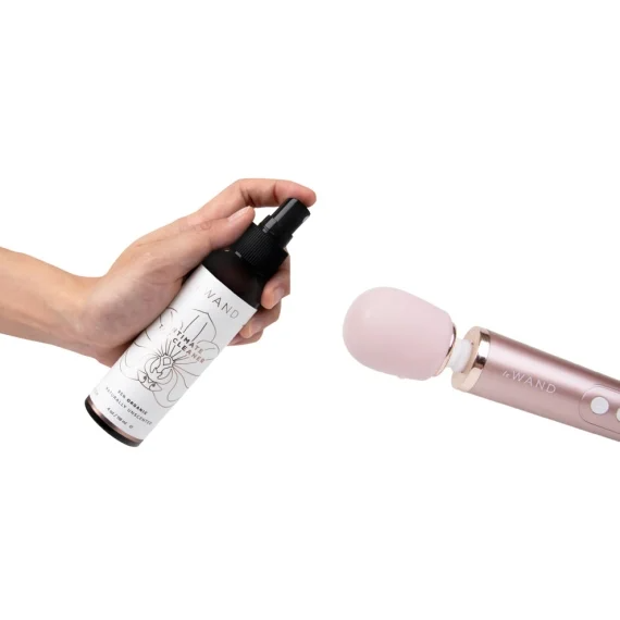 A hand holds a bottle of Le Wand intimate cleaner next to a pink Le Wand vibrator.