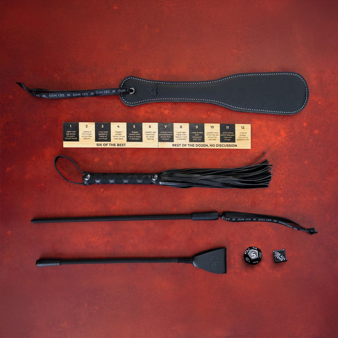 A 5-piece impact play toolkit with a paddle, flogger, crop, ruler, and dice.