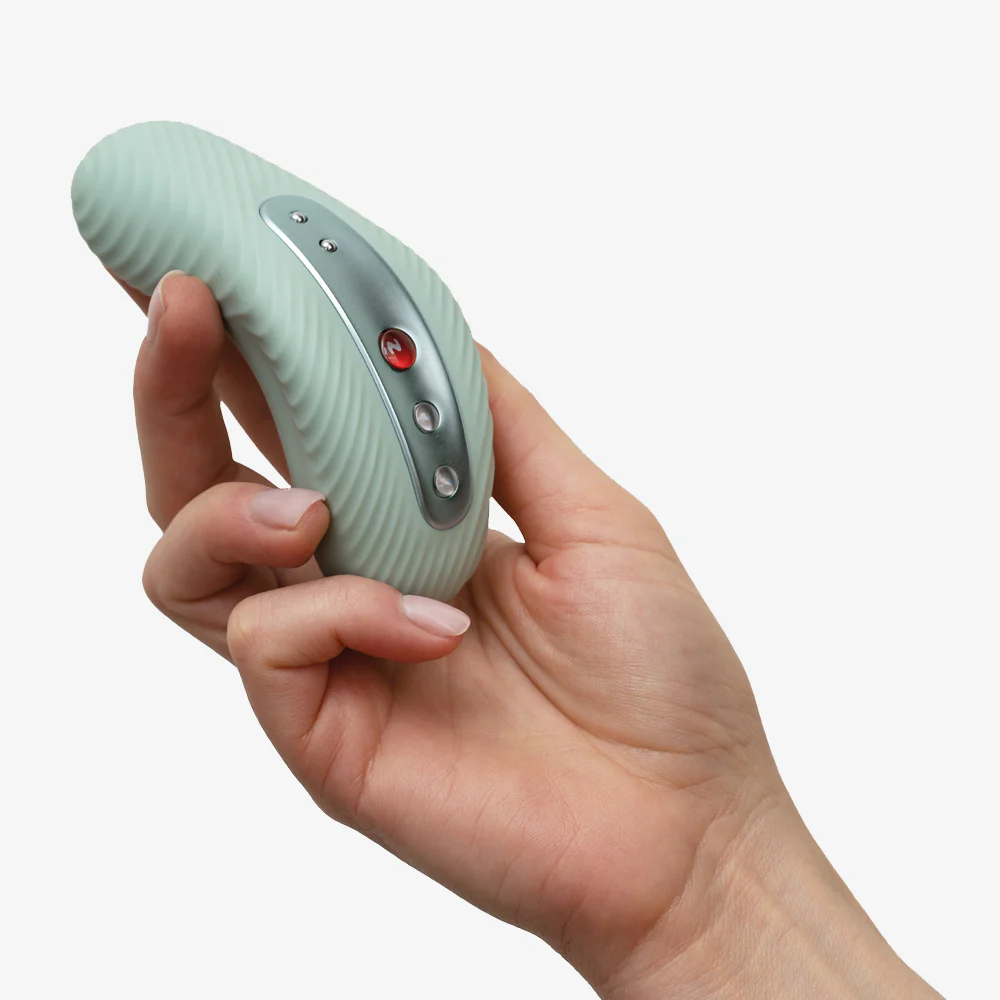 A hand holds a light green vibrating external massager with a silver control panel.