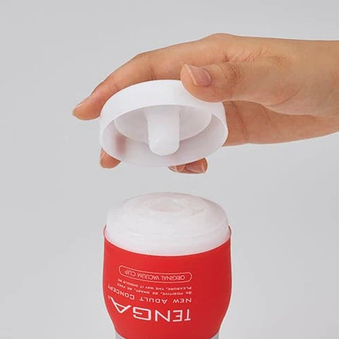 A hand holds the white cap of a red Tenga masturbator cup.