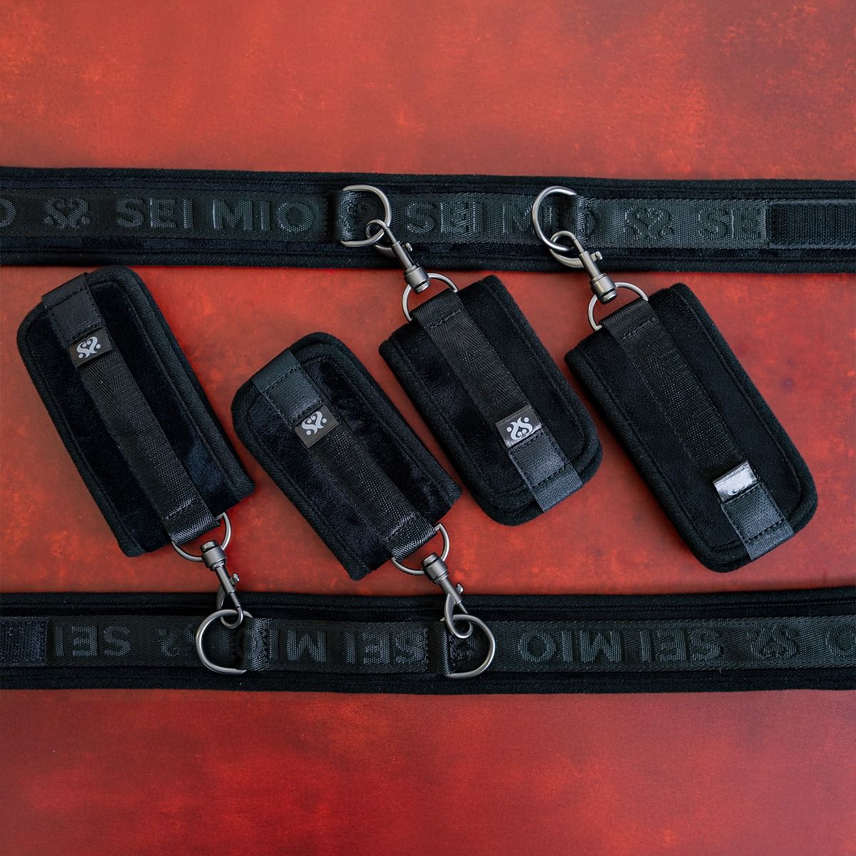 Black adjustable restraints with metal clips and a branded strap on a red background.
