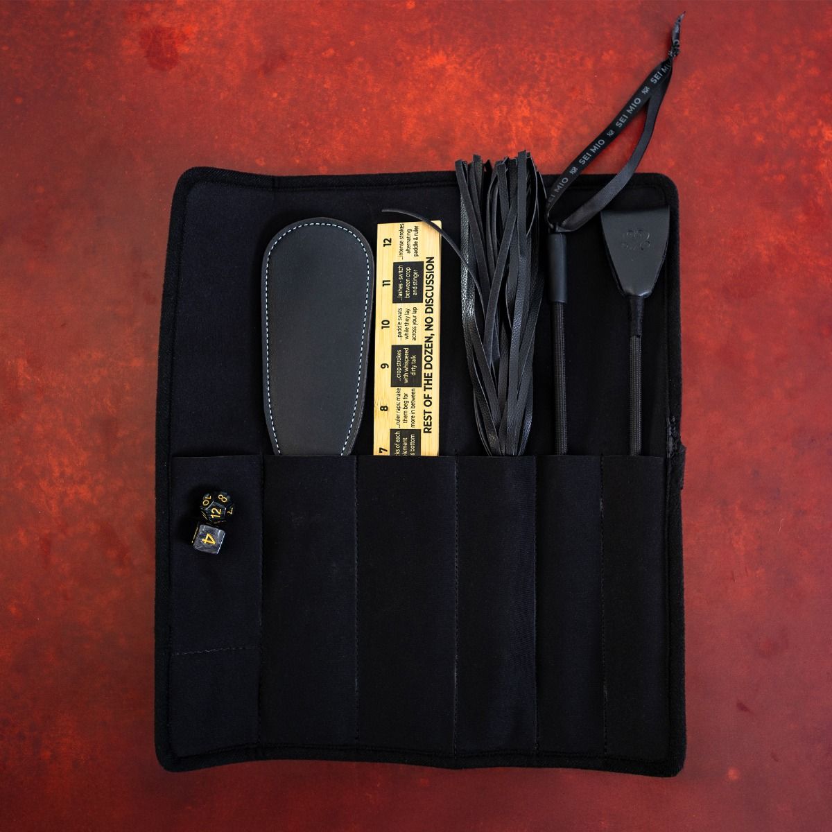 A black fabric roll-up case containing a paddle, flogger, crop, ruler, and dice.