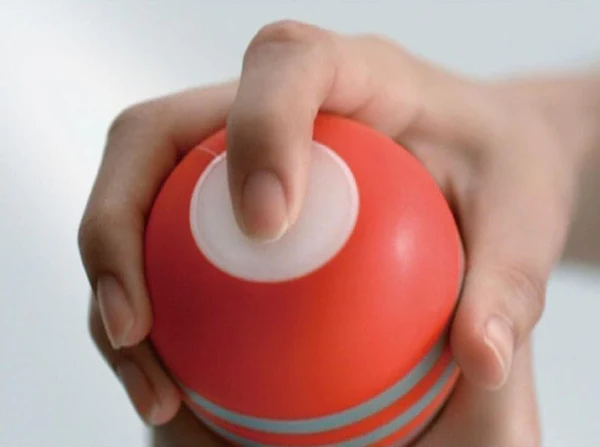 A hand holds a red Tenga Rolling Head Cup male masturbator with a white button.