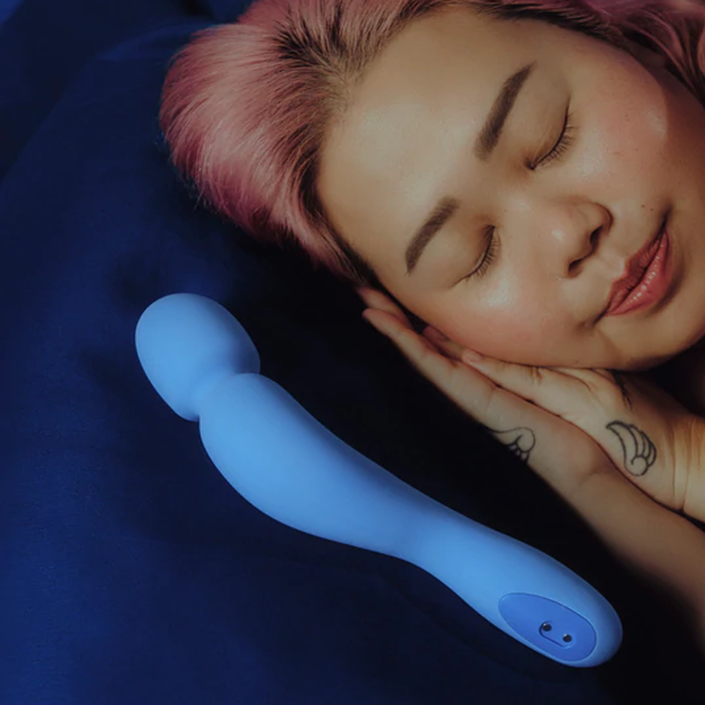 A periwinkle silicone wand vibrator rests on a dark blue surface next to a sleeping person.