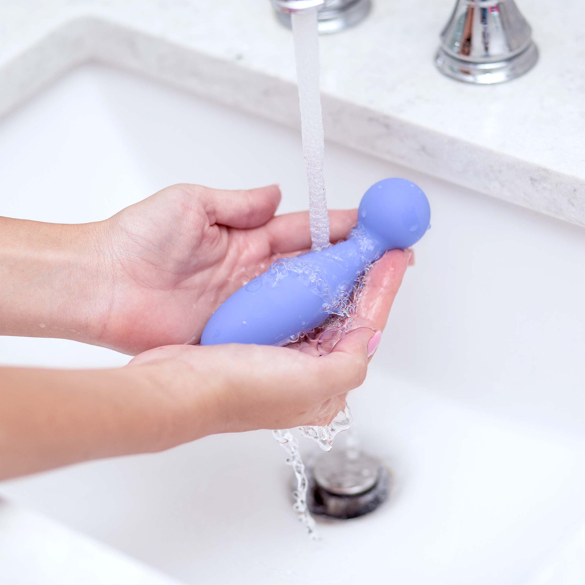 A person washes a purple vibrating personal massager under running water in a sink.