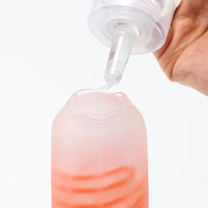 A hand squeezes a clear bottle, dispensing clear lubricant onto a pink, frosted bottle.