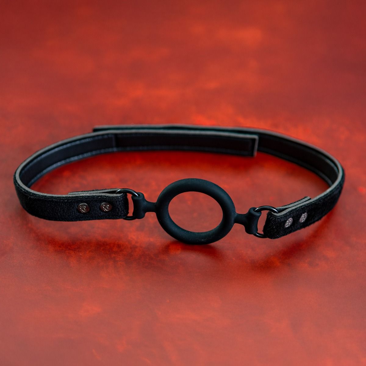Black O-ring gag with adjustable straps on a red textured background.