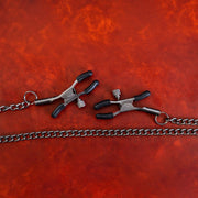 Two adjustable nipple clamps with black rubber tips are connected by a silver chain on a textured red surface.