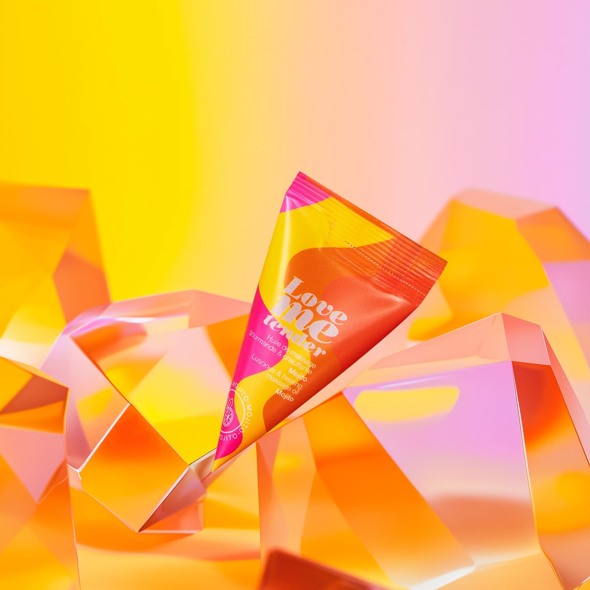 A tube of Love Me Tender Mojito warming massage oil rests on faceted orange and yellow crystals.