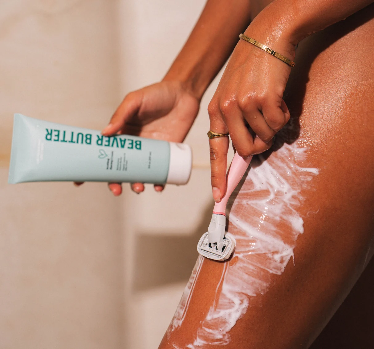 A hand holds a tube of Beaver Butter shave butter while shaving a leg with a pink razor.