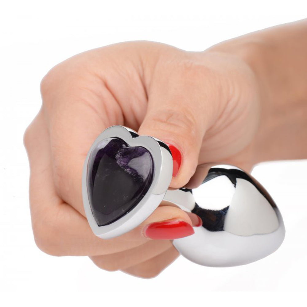 A hand holds a heart-shaped anal plug with an amethyst gemstone and a weighted aluminum base.