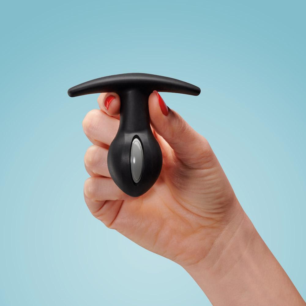 A hand holds a black weighted anal plug with a gray oval button.