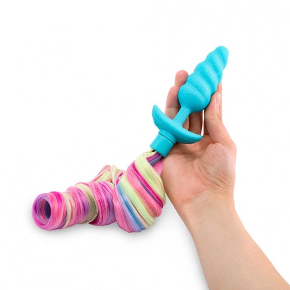 A hand holds a blue vibrating unicorn anal plug with a long, rainbow-colored tail.