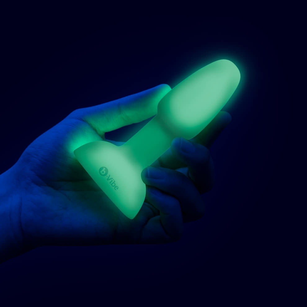A hand holds a glowing green anal plug with the b-Vibe logo.