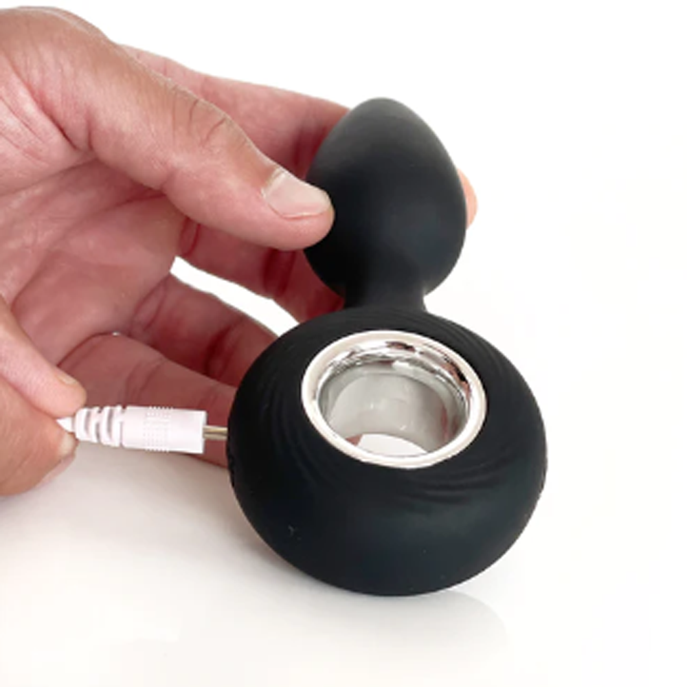 A hand holds a black vibrating anal plug with a silver ring and a white charging cable.