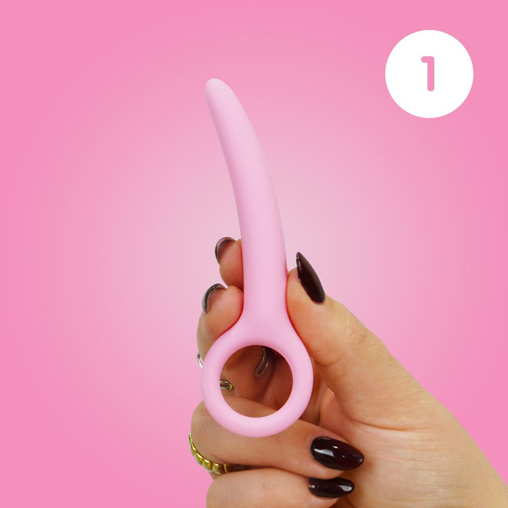 A pink curved silicone dilator with a finger loop is held by a hand.
