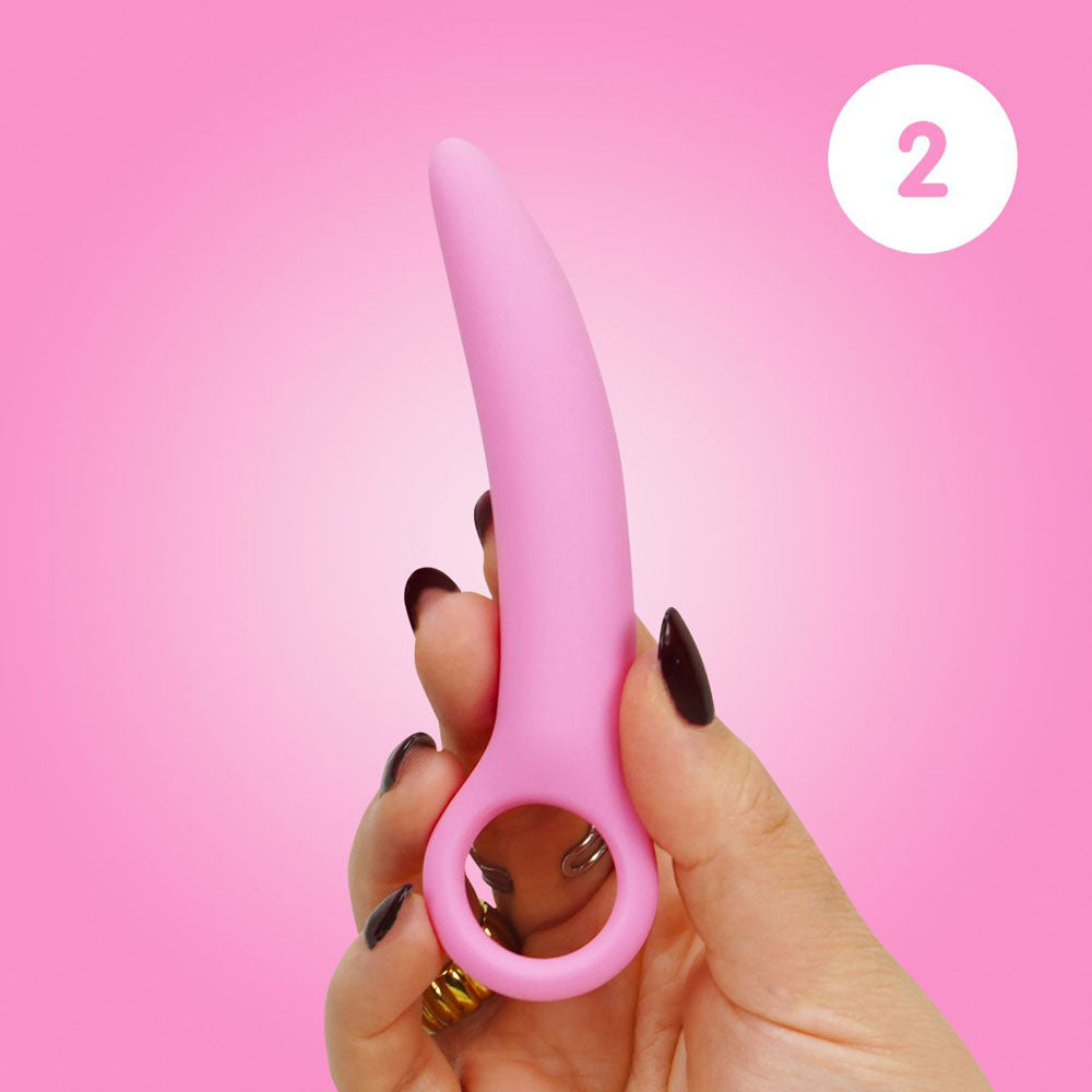 A hand holds a pink curved silicone dilator with a finger loop.