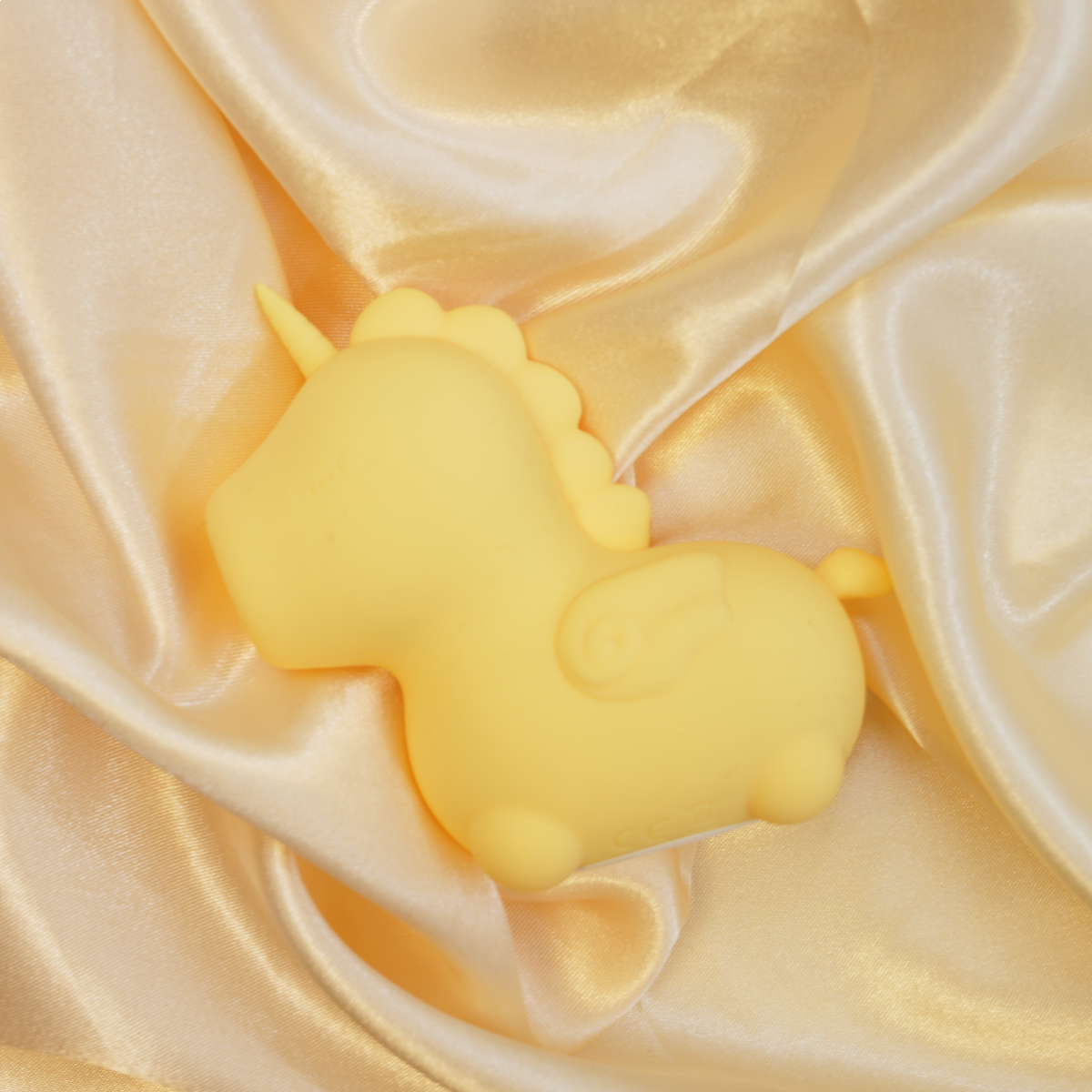 A yellow silicone unicorn toy with wings rests on satin fabric.