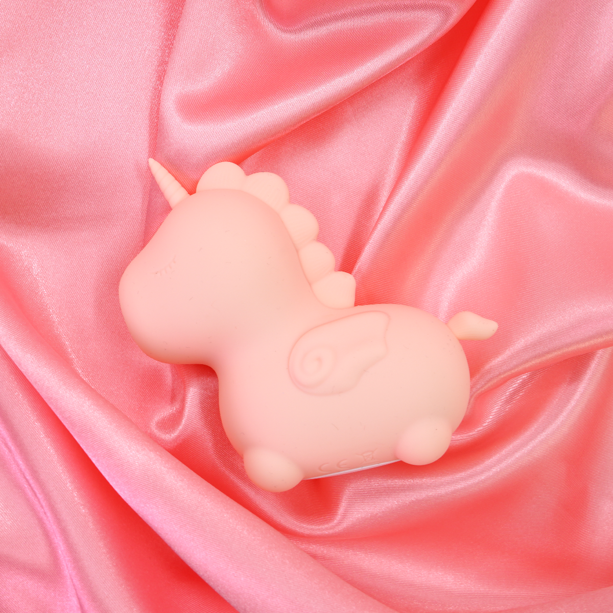 A pink silicone unicorn vibrator with wings rests on pink satin fabric.