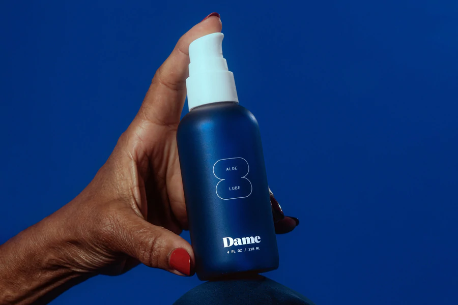 A hand holds a blue bottle of Dame Aloe Lube with a white pump.