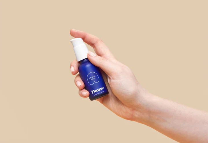 A hand holds a blue bottle of Dame Arousal Serum with a white pump.