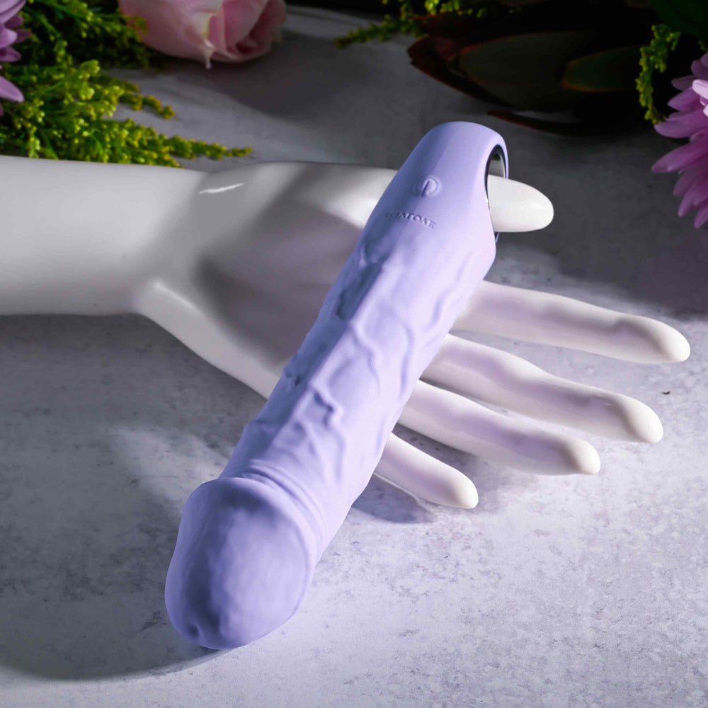 A purple textured vibrator rests on a white mannequin hand with flowers in the background.
