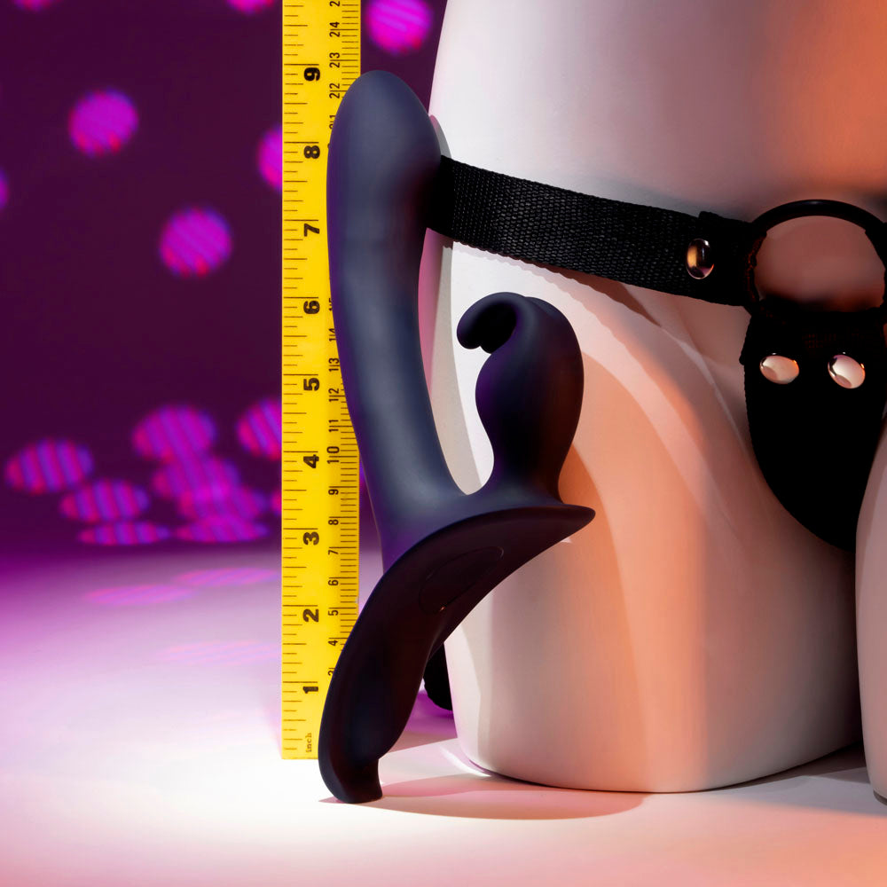 A dark purple strap-on rabbit vibrator with a curved shaft and ear stimulator is measured against a ruler.