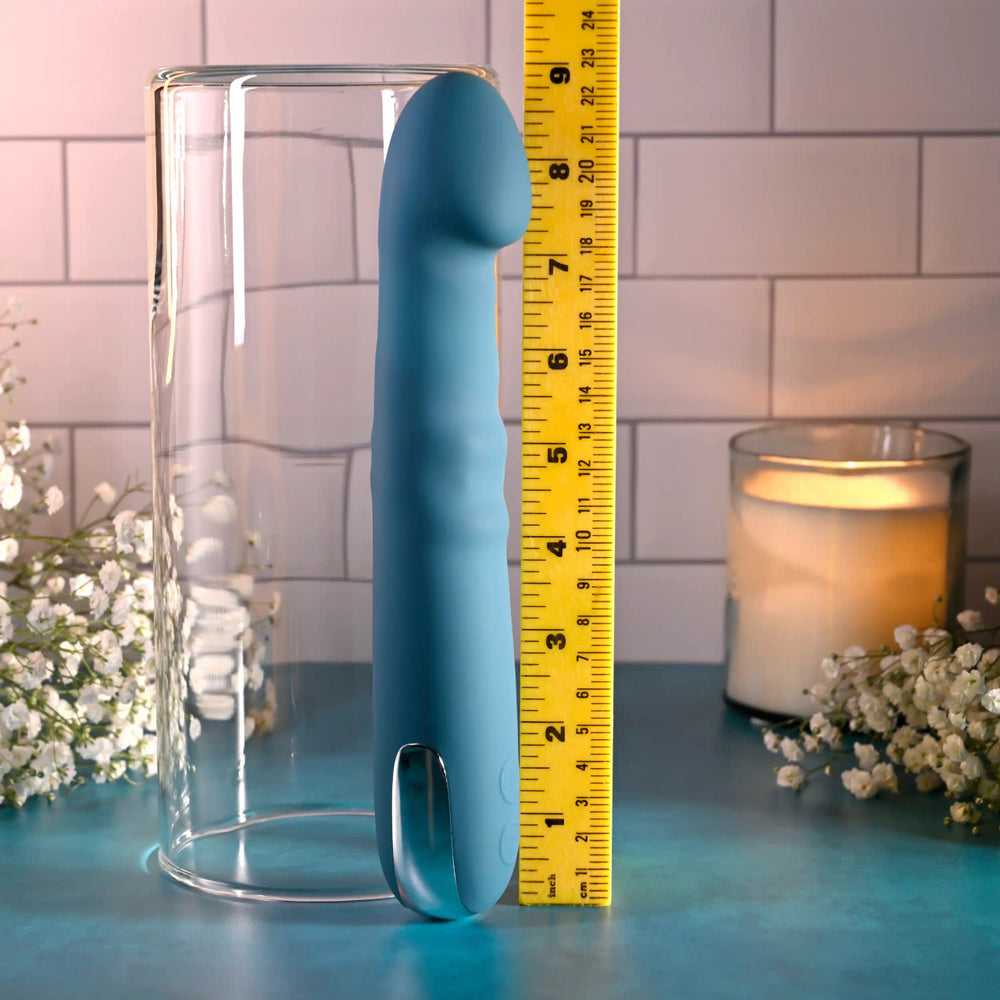 A blue silicone vibrator is measured against a ruler, next to a glass cylinder.