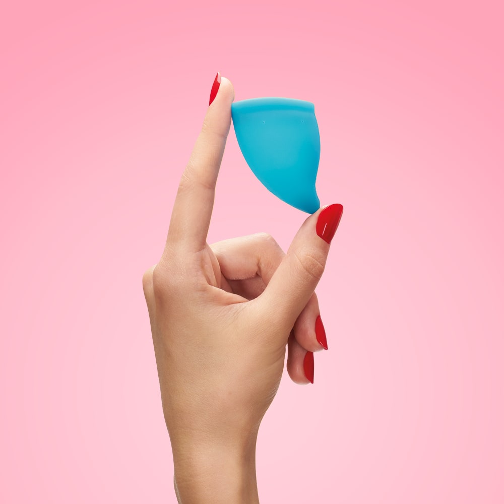 A hand holds a blue menstrual cup against a pink background.