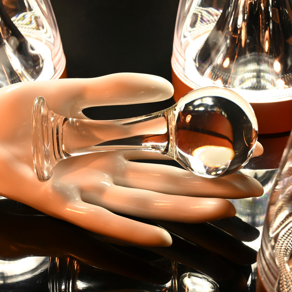 A glass ball anal plug with a flared base rests on a mannequin hand.