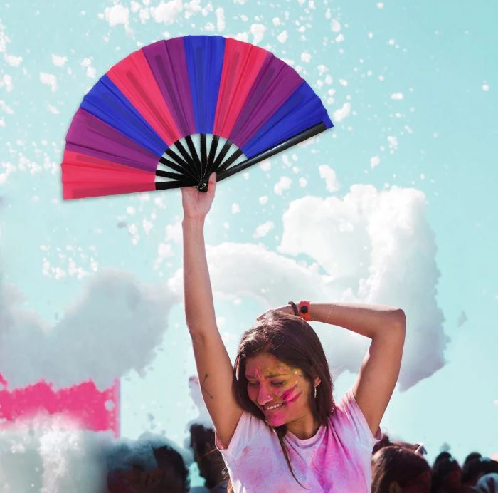A large folding hand fan with bisexual pride colors is held up by a person.