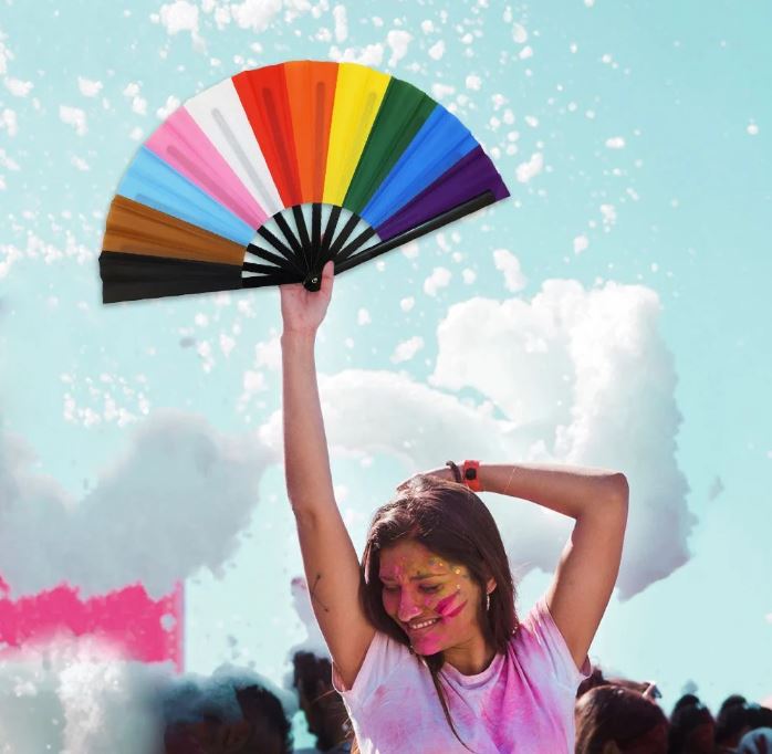 A Progress Pride flag folding hand fan held by a person with colorful powder on their face.