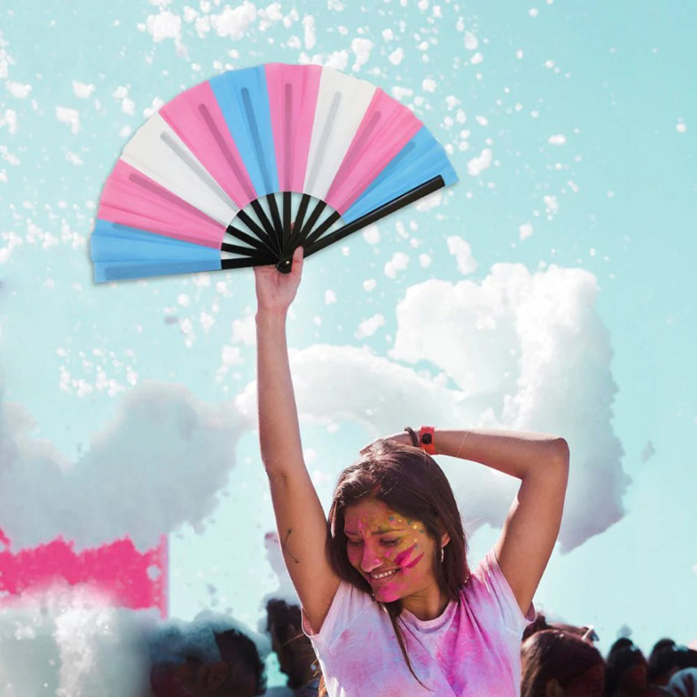 A large folding hand fan with transgender pride colors is held up against a blue sky.