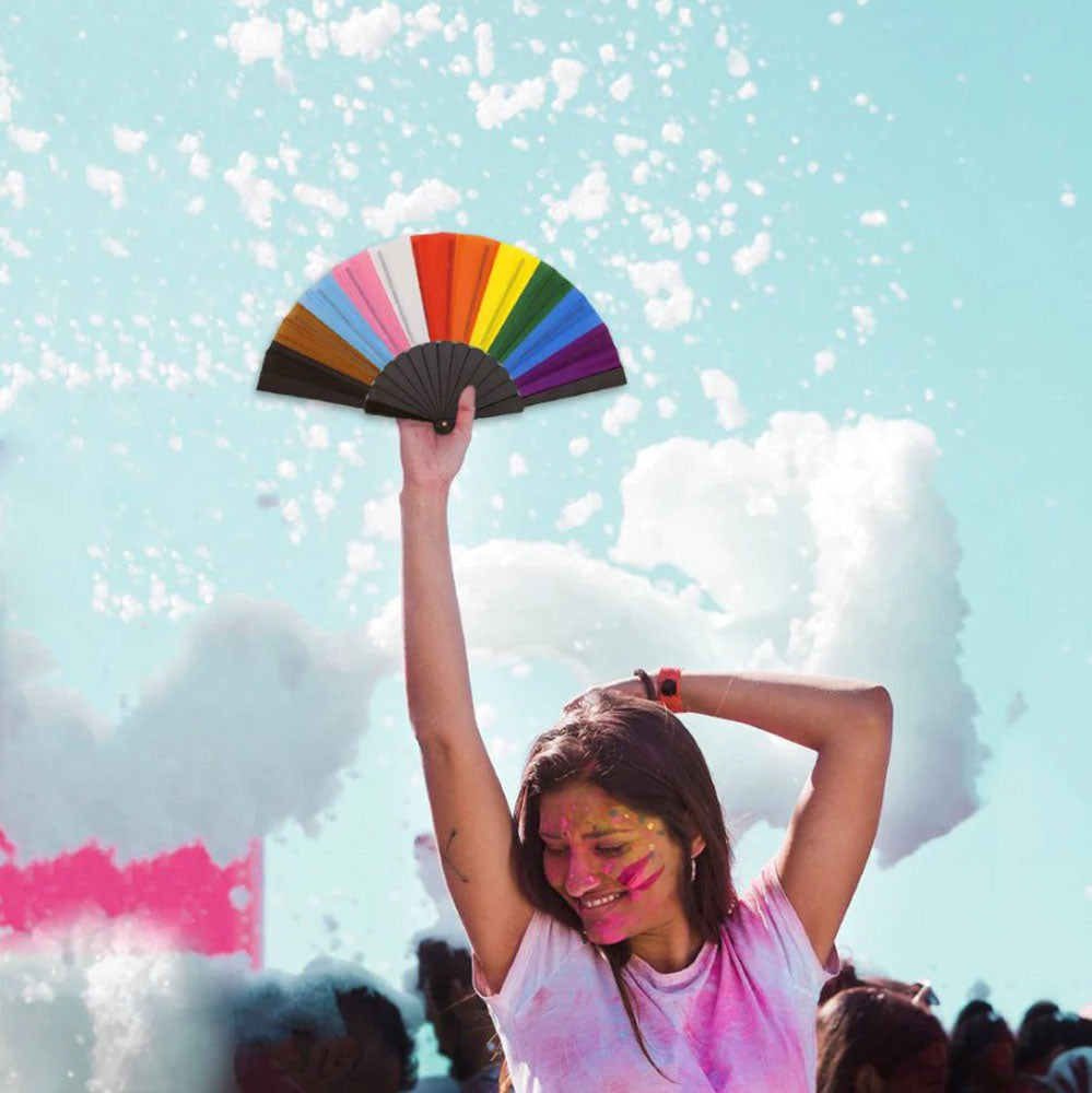 A hand holds a colorful Progress Pride fan against a bright sky.