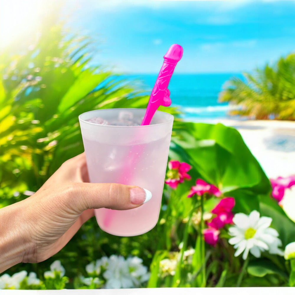 A hand holds a pink drink with a penis-shaped straw against a tropical beach background.
