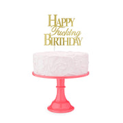 Gold glitter cake topper with "Happy Fucking Birthday" text on a pink cake stand.