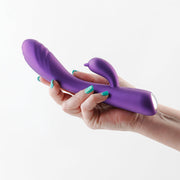 A purple silicone vibrator with a rabbit ear attachment held in a hand.