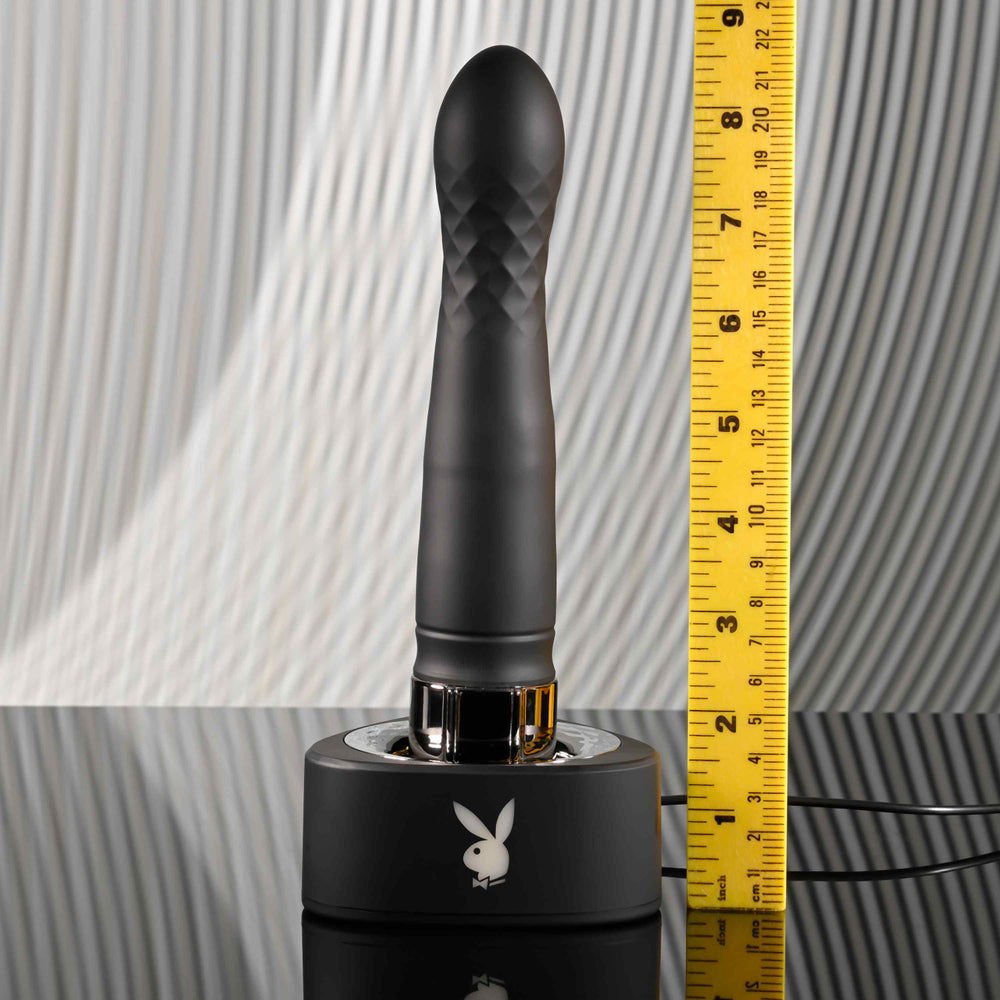 Black silicone vibrator with Playboy logo on charging base, next to a ruler.