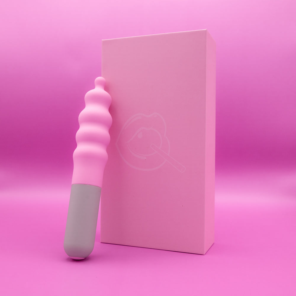 A pink and gray vibrator rests next to a pink box with a lip and lollipop graphic.