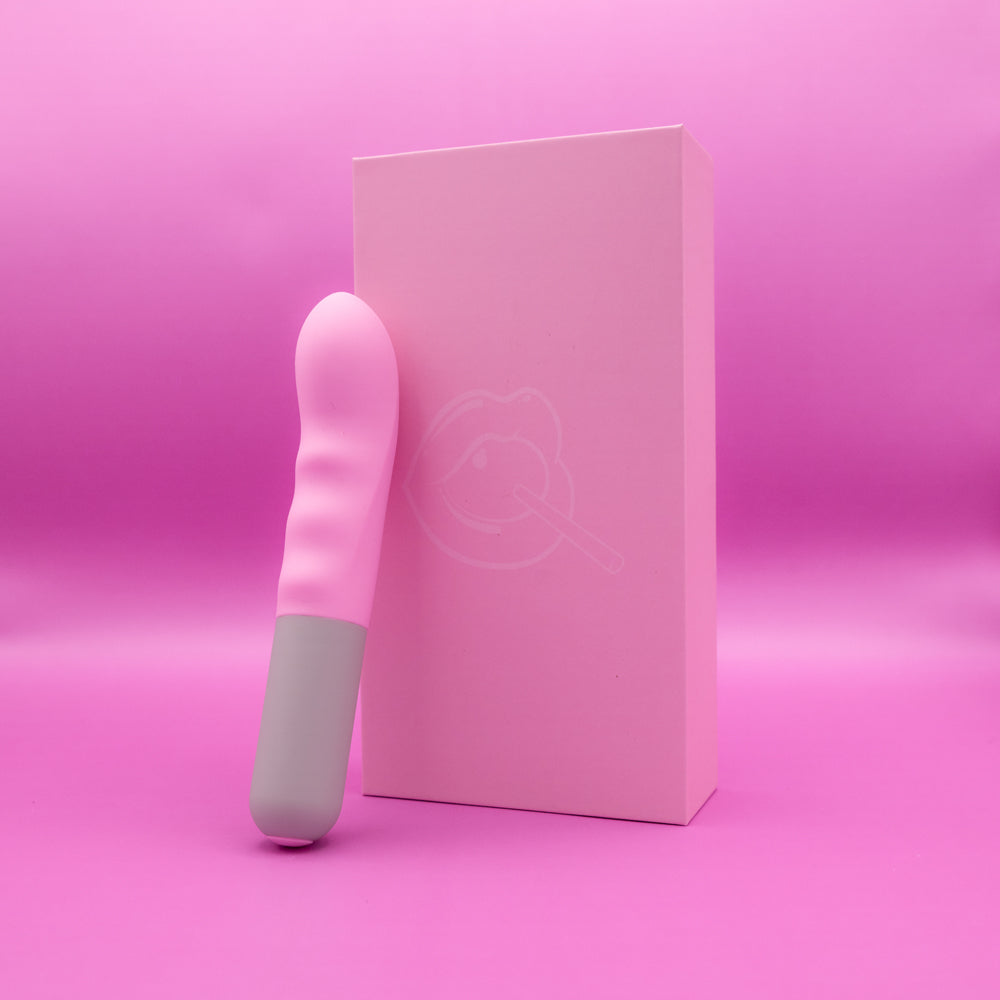 A pink and gray textured G-spot vibrator stands next to a pink box with a lip and lollipop graphic.