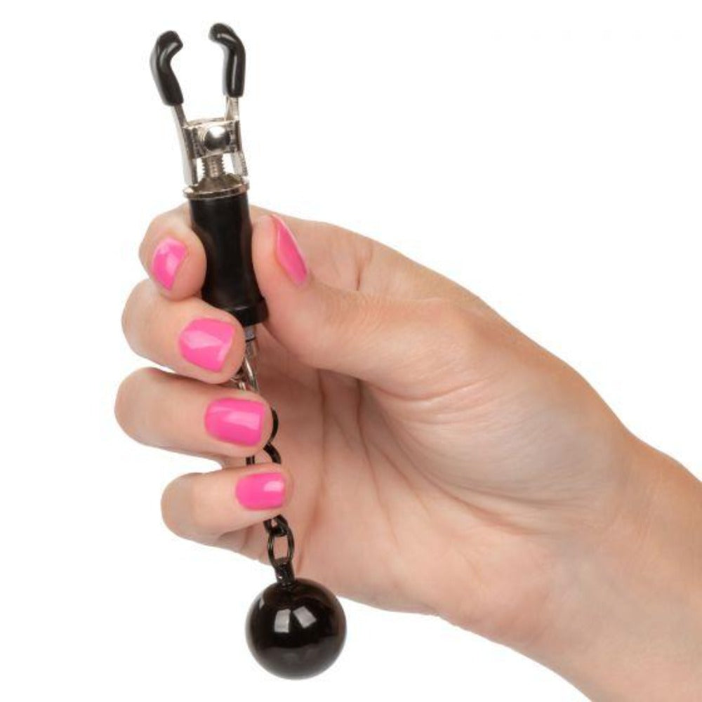 A hand holds weighted nipple clamps with a black ball and adjustable tension.